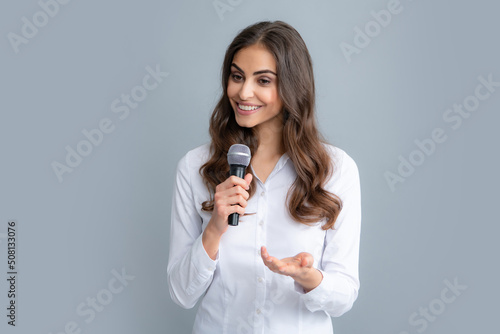 Beautiful business woman is speaking on conference. Happy smiling business woman holding mic, standing with microphone against grey background, wearing shirt.
