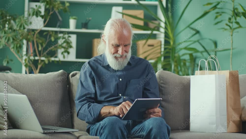 Progressive Senior Man is Sitting on the Sofa with Paper Box at Home and Shopping Online Using Tablet and Laptop.He is Cheerful for Good Purchases in the Network.Online Shopping and Retirement Concept Progressive Senior Man is Sitting on the Sofa with Paper Box at Home and Shopping Online Using Tablet and Laptop.He is Cheerful for Good Purchases in the Network.Online Shopping and Retirement Concept