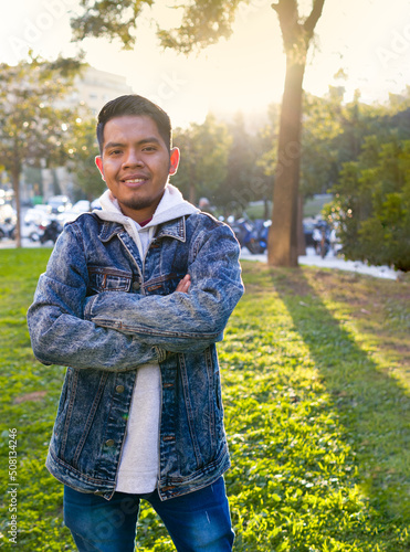 nice style brunette guy standing on a grass area with his arms crossed in a background of unfocused motor vehicles and the sunlights.