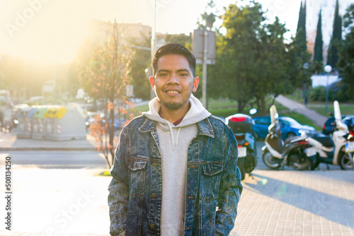 waist up shot of a originarian mexican feature man standing with hands in pockets on a urban place with land vehicles and the sun lights behind him