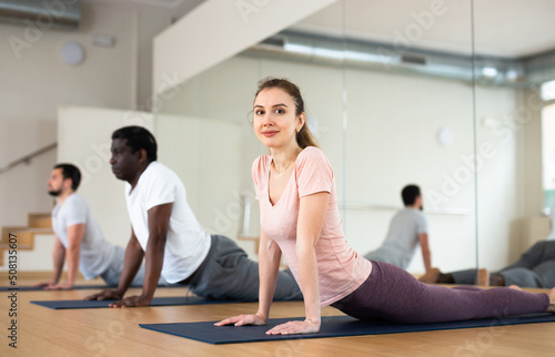 Young woman exercising cobra pose with group, yoga training in gym.