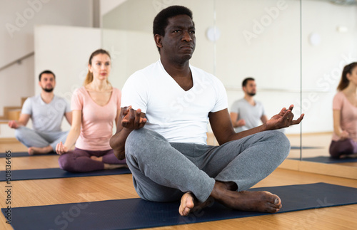 African-american man practising lotus pose with people during group yoga training.