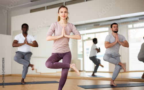 Motivated young attractive woman standing in balancing asana Eka Pada Utkatasana with hands clasped in prayer during group yoga class in fitness studio