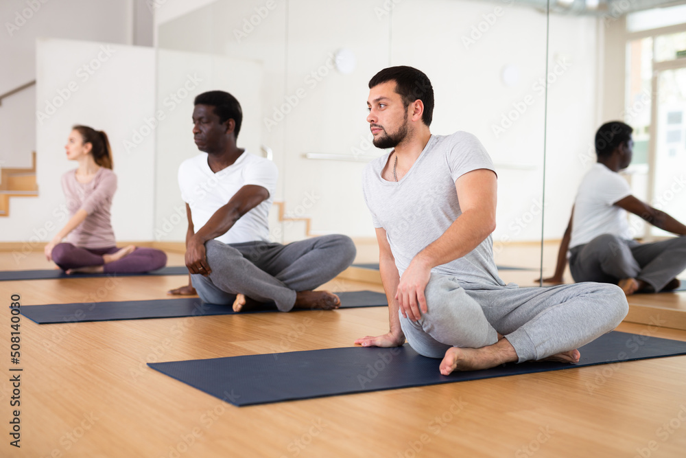 Fototapeta premium Young bearded man practicing Parivritta Sukhasana during group yoga training, sitting on mat in lotus position and doing side twist..