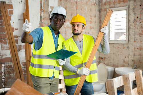 Two qualified builders, standing on a construction site in a room with a wooden bar, take notes on a tablet, evaluating ..the work plan photo