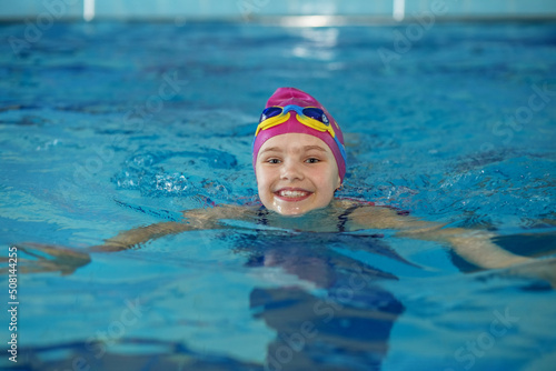 Happy child girl swims in swimming pool. Swim cap and goggles. Training and sports concept.