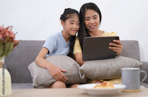 Happy mother and her cute daughter sitting in comfortable sofa together and using digital tablet. Weekend family leisure.