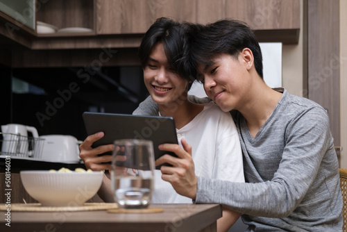 Cheerful gay couple embracing and using digital tablet together in their kitchen. LGBT and love concept.