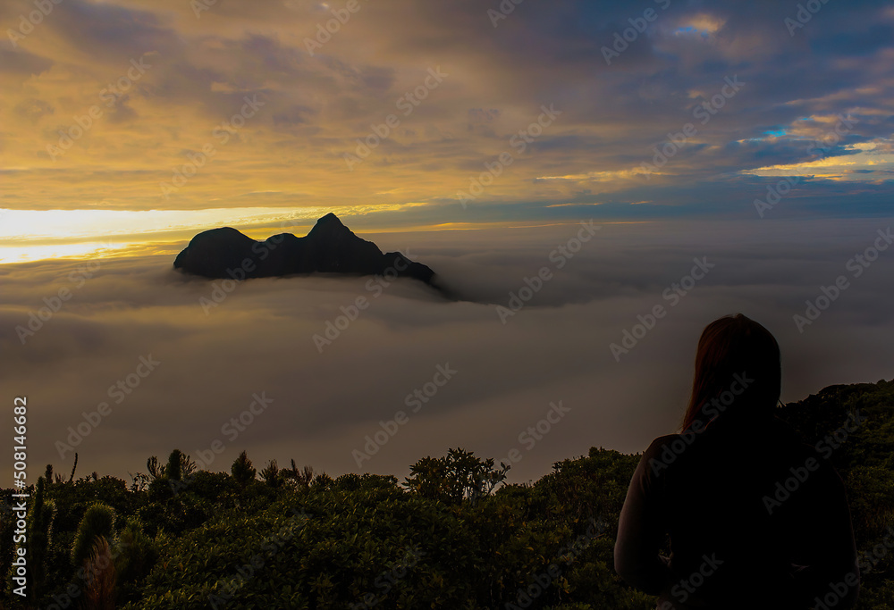 Pico Paraná, ponto mais alto do sul do Brasil, ao nascer do sol, em meio às nuvens, sendo observado por uma mulher, 