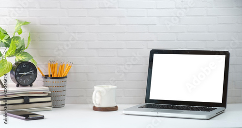Mock up computer laptop, alarm clock, coffee cup, books and houseplant on white table against brick wall.