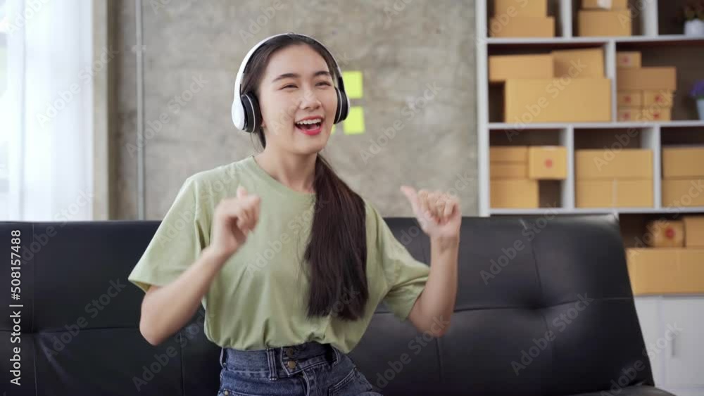 Attractive Asian woman is having fun at home listening to music through headphones holding smartphone, singing and dancing on sofa. Modern technology and young people concept. Attractive Asian woman is having fun at home listening to music through headphones holding smartphone, singing and dancing on sofa. Modern technology and young people concept.