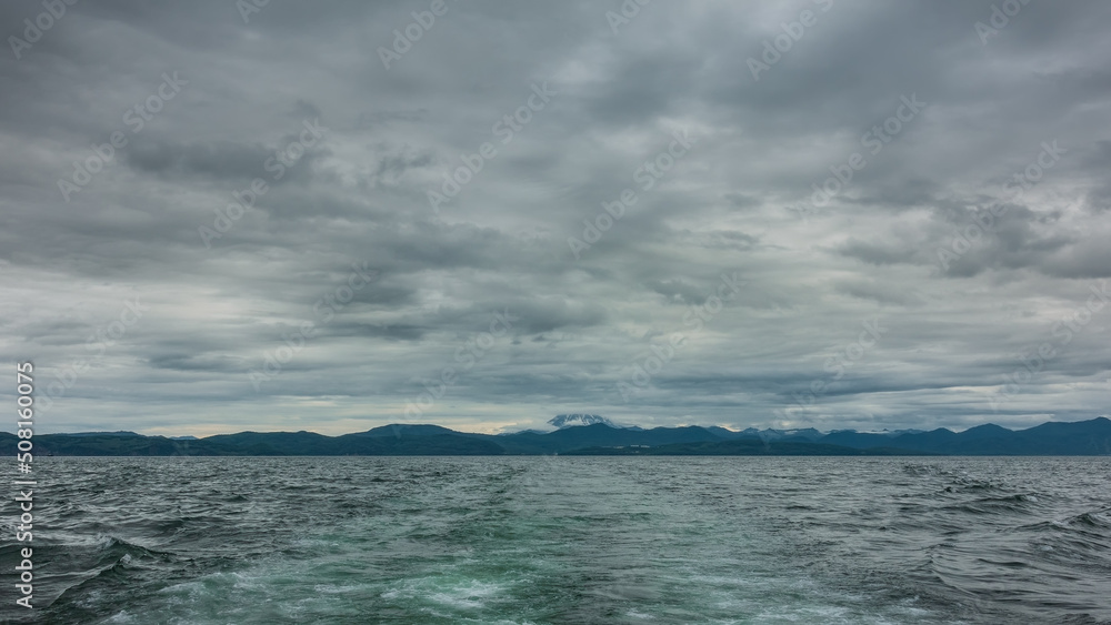 The foam trail from the boat is visible on the turquoise water of the Pacific Ocean. Silhouettes of coastal hills in the distance. The volcano is hidden in dense clouds. Kamchatka. Avacha Bay