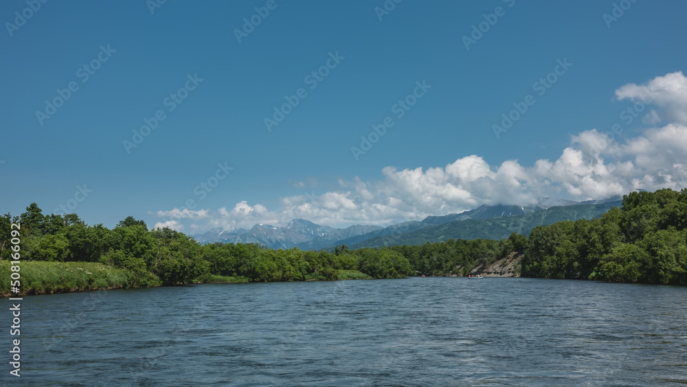 A calm blue river flows under an azure sky. A rafting boat is visible in the distance. Lush green vegetation on the banks. A mountain range against the background of clouds. Kamchatka