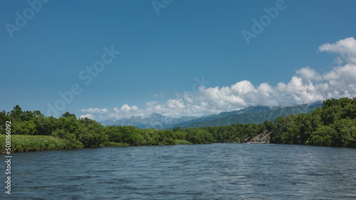 A calm blue river flows under an azure sky. A rafting boat is visible in the distance. Lush green vegetation on the banks. A mountain range against the background of clouds. Kamchatka © Вера 