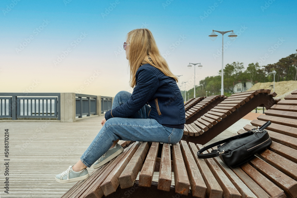 A young lonely blonde woman on an empty city embankment rests sitting on a sun lounger on a spring or autumn evening and looks into the distance. The girl misses the warmth and summer. A young lonely blonde woman on an empty city embankment rests sitting on a sun lounger on a spring or autumn evening and looks into the distance. The girl misses the warmth and summer.