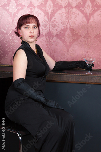 sensetive lass wearing black robe and long black gloves is sitting on a bar chair with glass of alcoholic beverages in her hand photo