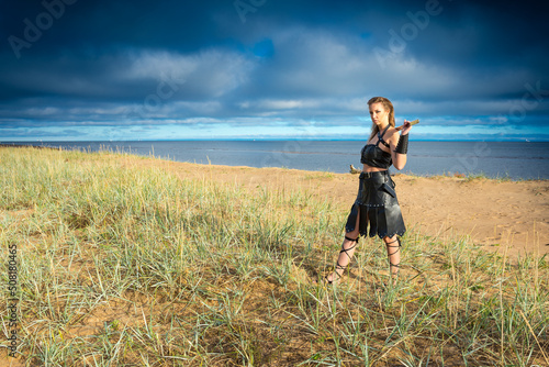 Middle-Age battler woman wearing dark leather armour with iron morglay in her hands is standing on the sandy beach photo