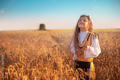 Young girl with traditional Bulgarian folklore costume at the agricultural wheat field during harvest time with industrial combine machine
