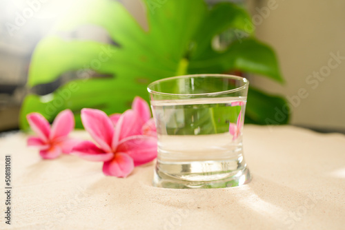 Glass Mineral Water On Sand Next To Pink Frangipani Flowers.