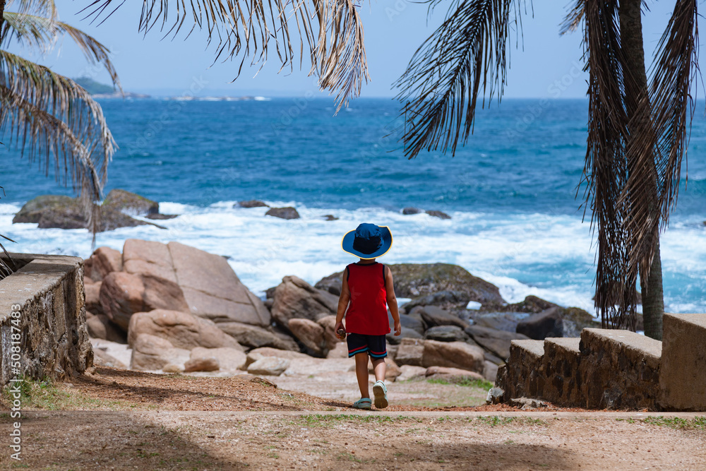 Little boy walking to a beautiful blue ocean with a hat
