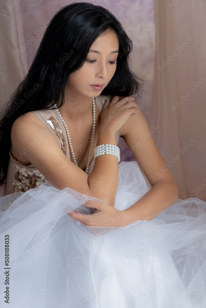 A beautiful Asian woman wearing a gold sequin top and tulle skirt with pearl jewellery and surrounded by draped fabric A beautiful Asian woman wearing a gold sequin top and tulle skirt with pearl jewellery and surrounded by draped fabric