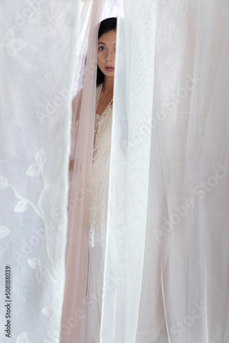 Beautiful Asian woman with long hair wearing a white lace dress and pearl jewellery surrounded by soft draped fabric