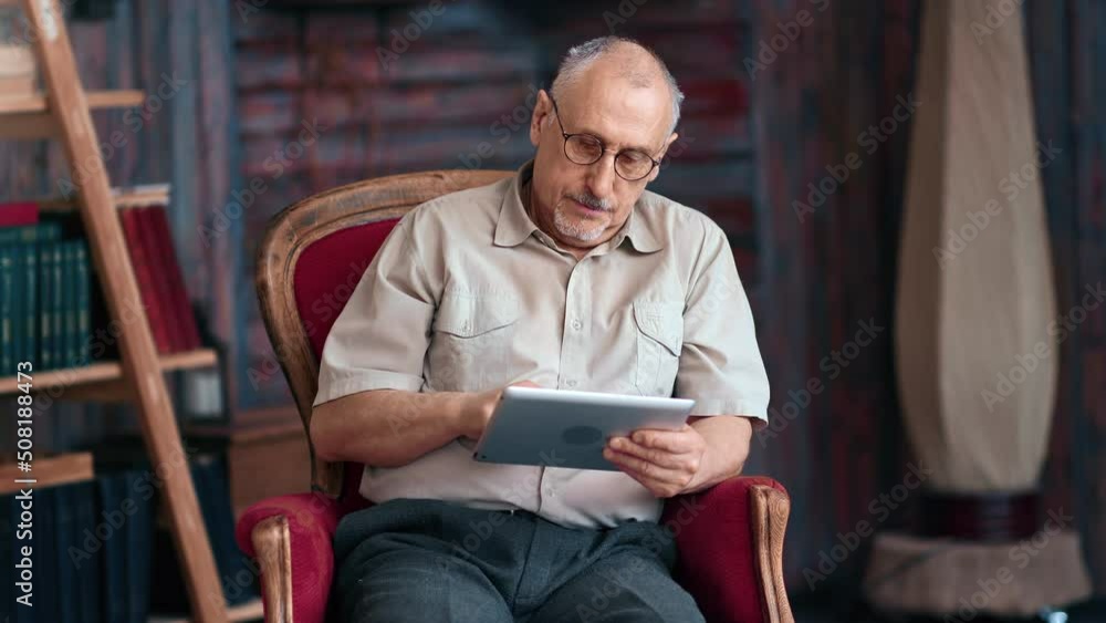 Elderly man browsing internet use tablet pc sitting on vintage armchair at rustic house interior Elderly man browsing internet use tablet pc sitting on vintage armchair at rustic house interior