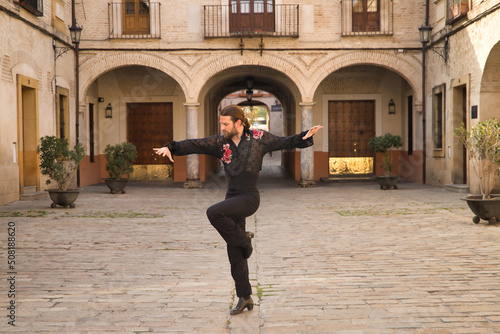 Young man with beard and ponytail, wearing black transparent shirt with black polka dots and red roses, black pants and jacket, dancing flamenco in the city. Concept art, dance, culture, tradition.