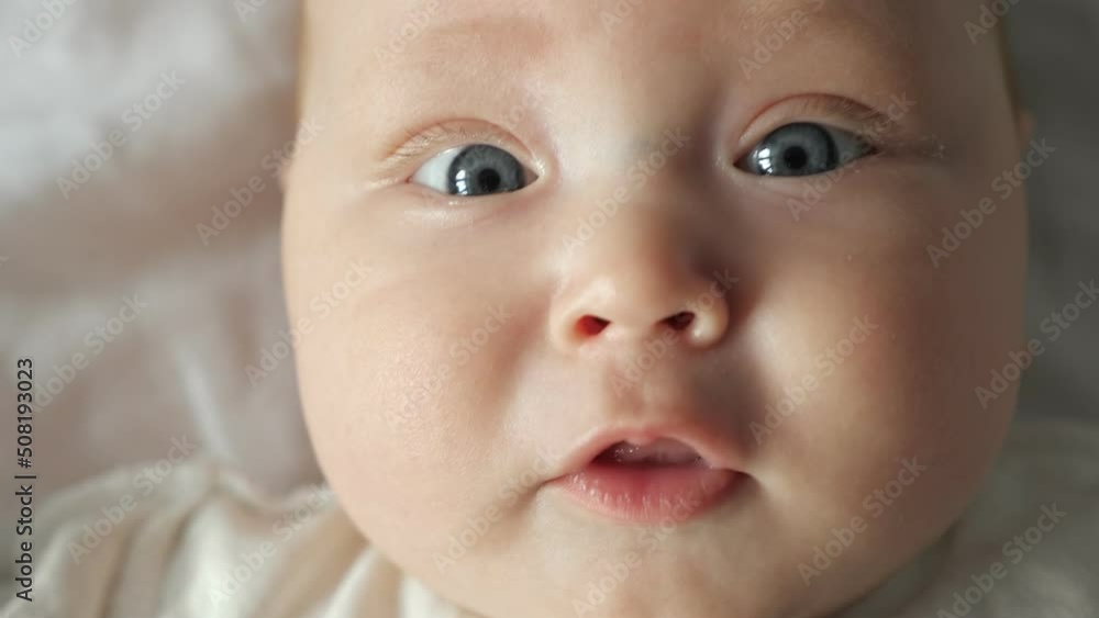 Cute face of little baby girl with large light grey eyes. Newborn lies on white sheets and wiggles arms with cheerful face expression extreme closeup Cute face of little baby girl with large light grey eyes. Newborn lies on white sheets and wiggles arms with cheerful face expression extreme closeup