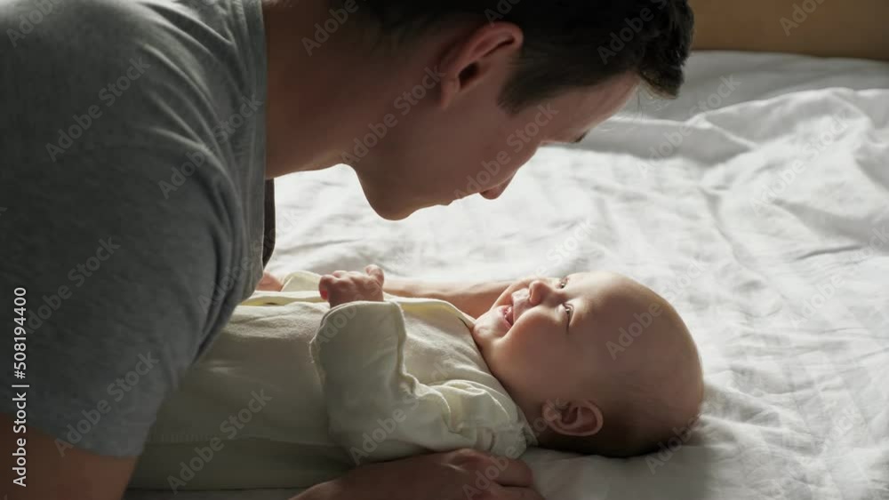 Young happy father tries to speak and play with newborn daughter. Cheerful little baby girl lies on bed with white sheets and looks at father closeup Young happy father tries to speak and play with newborn daughter. Cheerful little baby girl lies on bed with white sheets and looks at father closeup