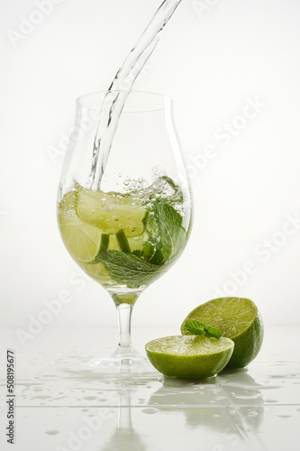 Clear shiny glass filled with with lime and mint and delicious Mojito cocktail composed on a white background. Pouring water into glass. Shallow depth of field