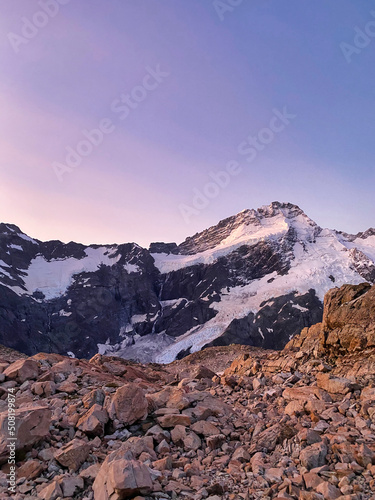 Pastel-colored picture of the mountain range around Mount Cook, New Zealand, with the sky turning purple during sunset and big heavy and rough rocks and gravel in the foreground.  © Philipp