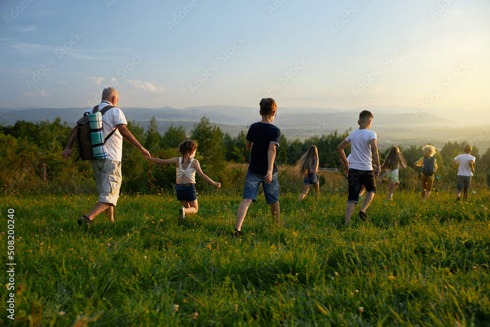 Fototapeta premium Back view of kids with old man leading each other forward in hills. Male with gray hair wearing rucksack together with kids hiking, traveling, running ahead. Concep of active rest.