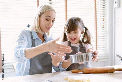 Happy Caucasian family enjoying spend time together in the kitchen. Beautiful mom is caring and teaching adorable daughter helping sprinkling dough with flour to make a bakery in kitchen at home.