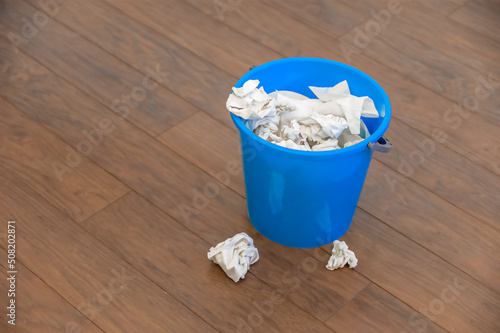 a blue plastic bucket filled with used tissues sits on a wooden floor.