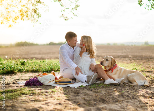 Portrait of a beautiful caucasian couple sitting on a blanket on a picnic and hugging and eating strawberries with a Labrador dog of sunset 
