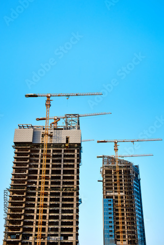 Two modern skyscrapers under construction with four yellow tower cranes along them in the process of installing windows on concrete structures against a clear blue sky in sunny weather