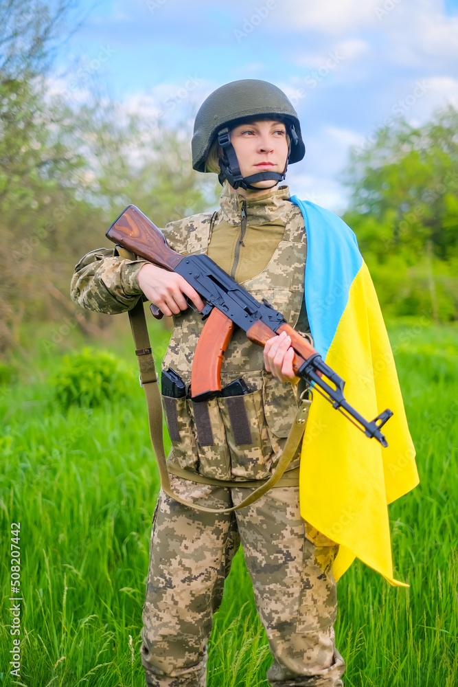 Armed with assault rifle determined female soldier in camouflage uniform and helmet, covered with Ukrainian flag shoulder looking away. Ukrainian woman patriot. Vertical orientation Armed with assault rifle determined female soldier in camouflage uniform and helmet, covered with Ukrainian flag shoulder looking away. Ukrainian woman patriot. Vertical orientation