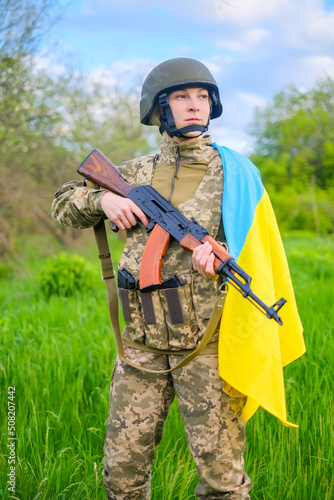 Armed with assault rifle determined female soldier in camouflage uniform and helmet, covered with Ukrainian flag shoulder looking away. Ukrainian woman patriot. Vertical orientation