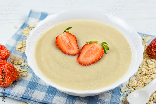 Oatmeal porridge for a baby with strawberries in a white bowl on a blue cloth napkin close-up. The first complementary food of a child, baby nutrition.