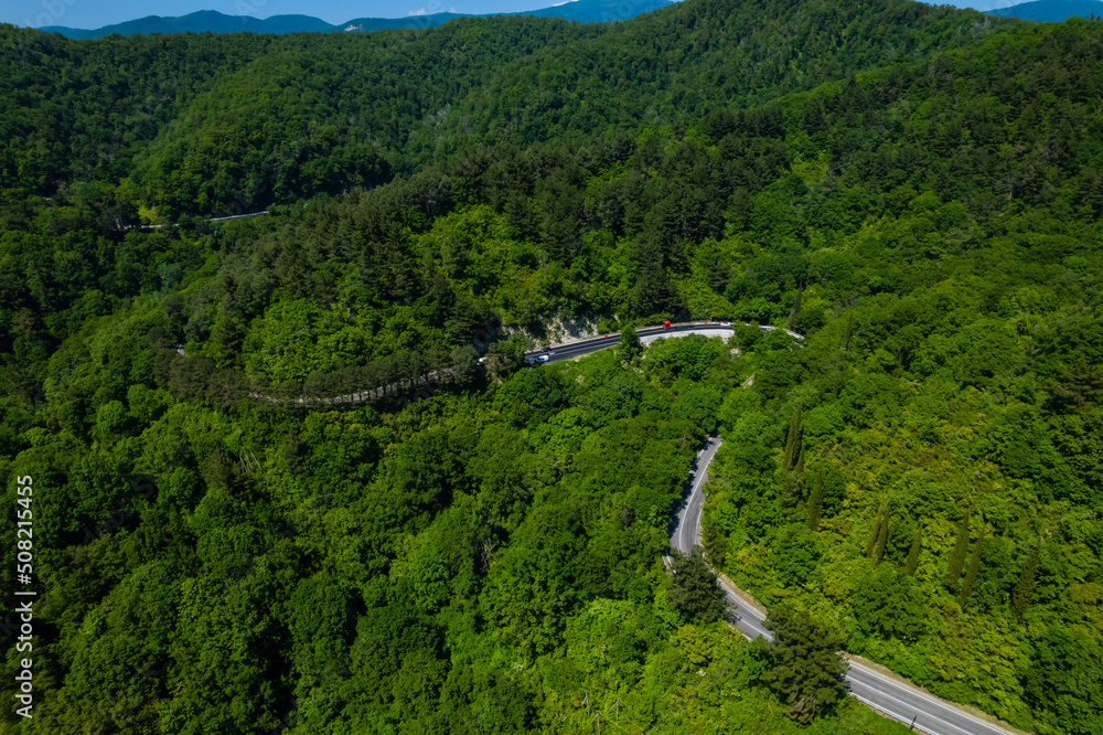 Asphalt road is meandering between blue sea and green mountains. Aerial view of car driving along the winding mountain road in Sochi, Russia. Asphalt road is meandering between blue sea and green mountains. Aerial view of car driving along the winding mountain road in Sochi, Russia.