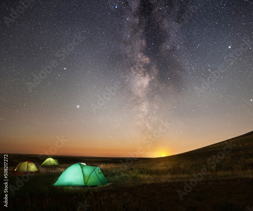 Travel and camping concept - three glowing camping tents at night under a starry sky with milky way and setting sun. Illuminated blue camping tent under stars at night