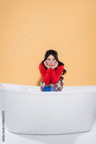 Stylish asian model sitting on bathtub on orange background.