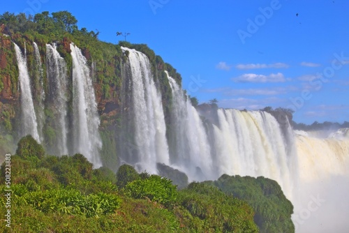 Foz do Igua  u Falls in Brazil. Bulky water continuously falling. Fog in the air and vision blurred by fog. International tourism  UN preservation area. Clear blue sky and green vegetation at the bott