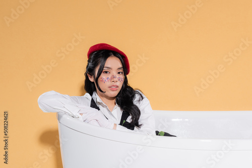 Stylish asian woman in gloves and beret sitting in bathtub on orange background.