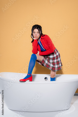 Stylish asian model posing in bathtub on orange background.