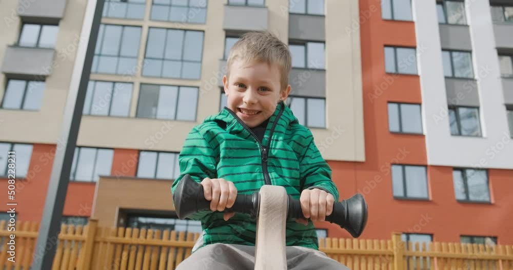 Small child riding toy horse swing, playing on playground. Kids activity outdoors, smiling and laughing boy looking in camera. Apartment residential area on background, family bought new flat Small child riding toy horse swing, playing on playground. Kids activity outdoors, smiling and laughing boy looking in camera. Apartment residential area on background, family bought new flat