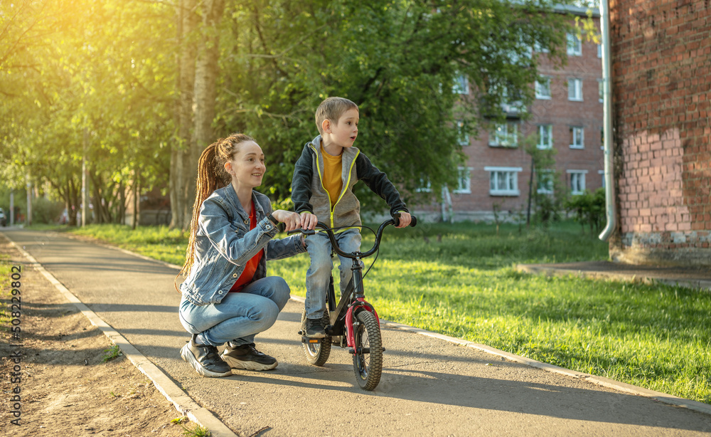 Mom helps a child boy learn to ride a two-wheeled bicycle in the park. A pleasant children's summer sports vacation