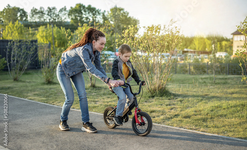 Mom helps a child boy learn to ride a two-wheeled bicycle in the park. A pleasant children's summer sports vacation