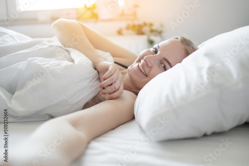 Positive blonde 30s woman with pillow lying in bed and smiling to camera at home. 